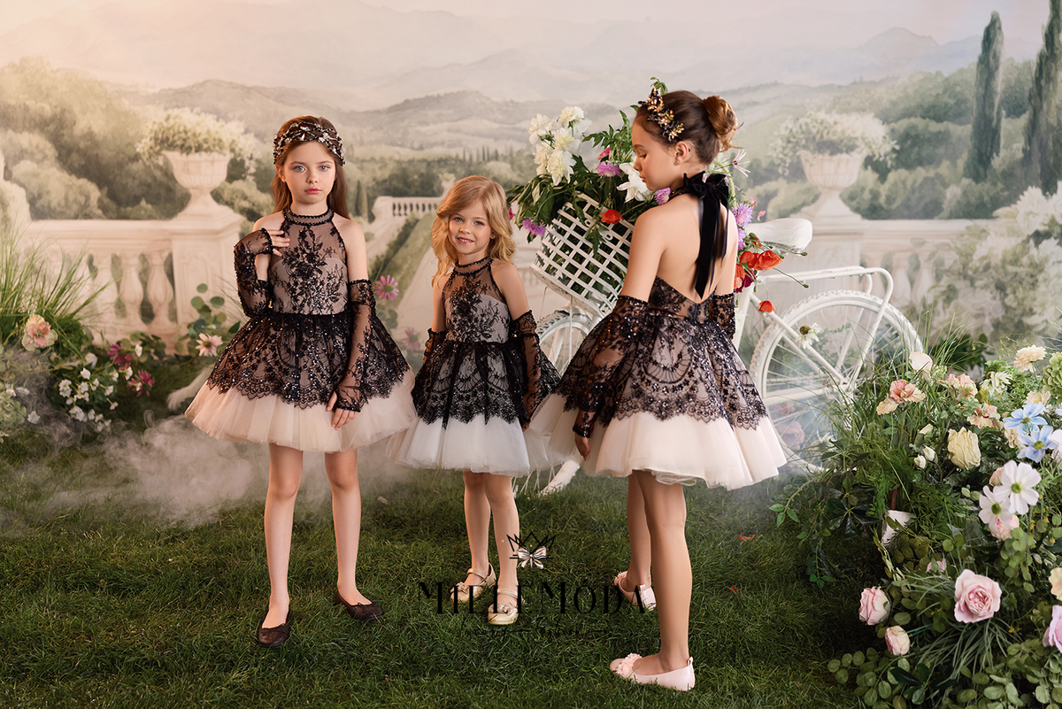 Three young girls wearing matching black lace and white tulle dress outfits pose together in a garden-themed studio setup with flowers, a white bicycle prop, and a scenic backdrop.