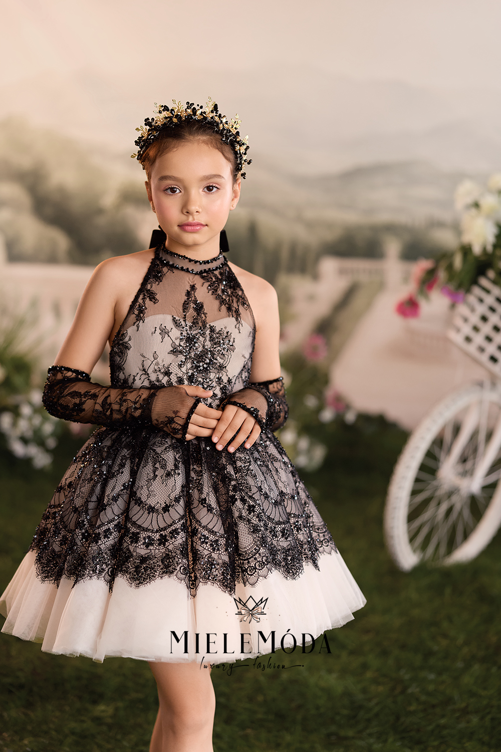 A young girl wearing a MieleMóda black lace and white tulle flower girl dress with matching long black lace sleeves and a floral crown, posed in front of a blurred garden background with a white carriage prop.