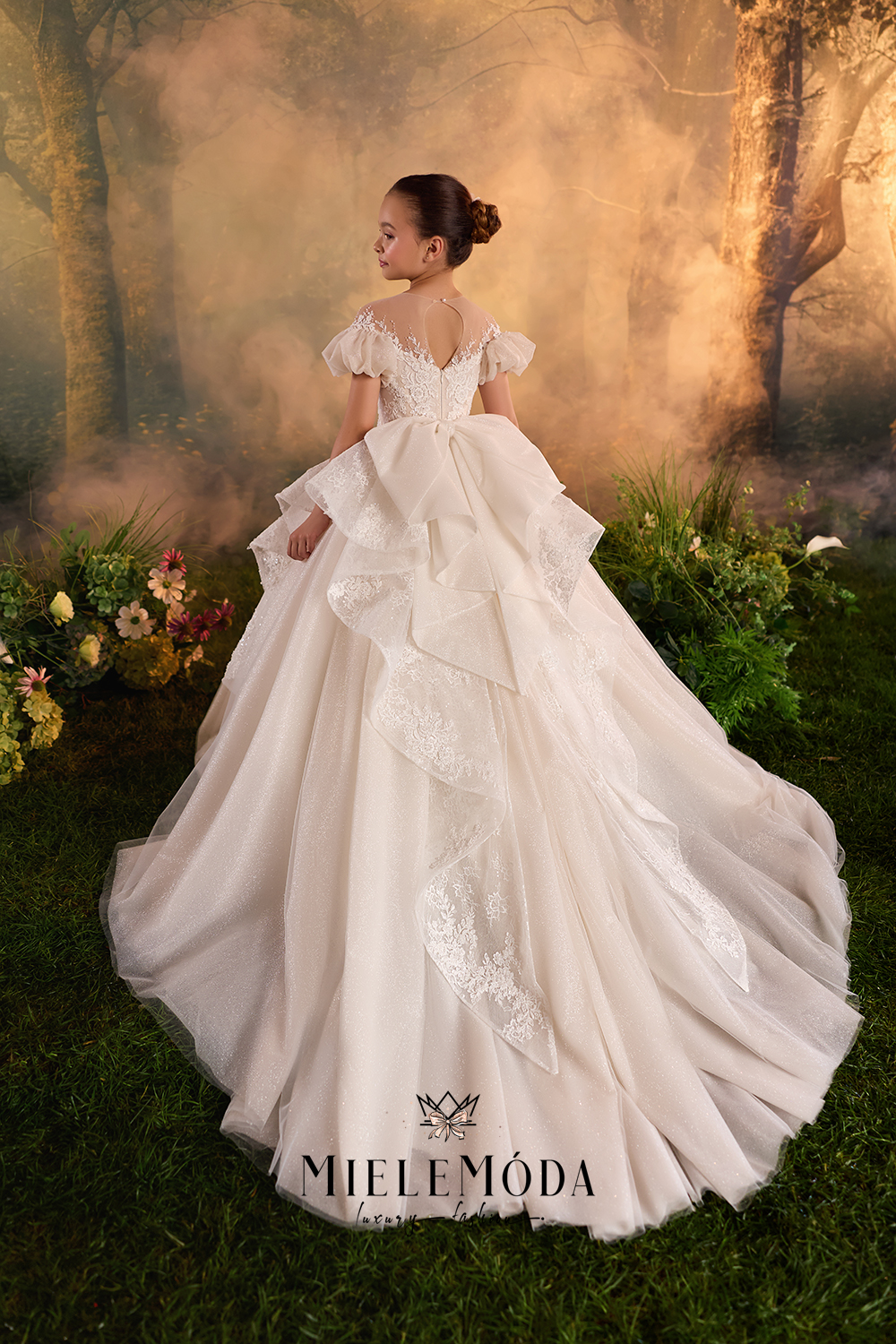 A young girl wearing a Miele Móda ball gown, photographed from behind in a fantasy forest setting with mist and flowers. The dress is off-the-shoulder with a ruffled bow at the back of the neck and a layered tulle skirt with lace details.
