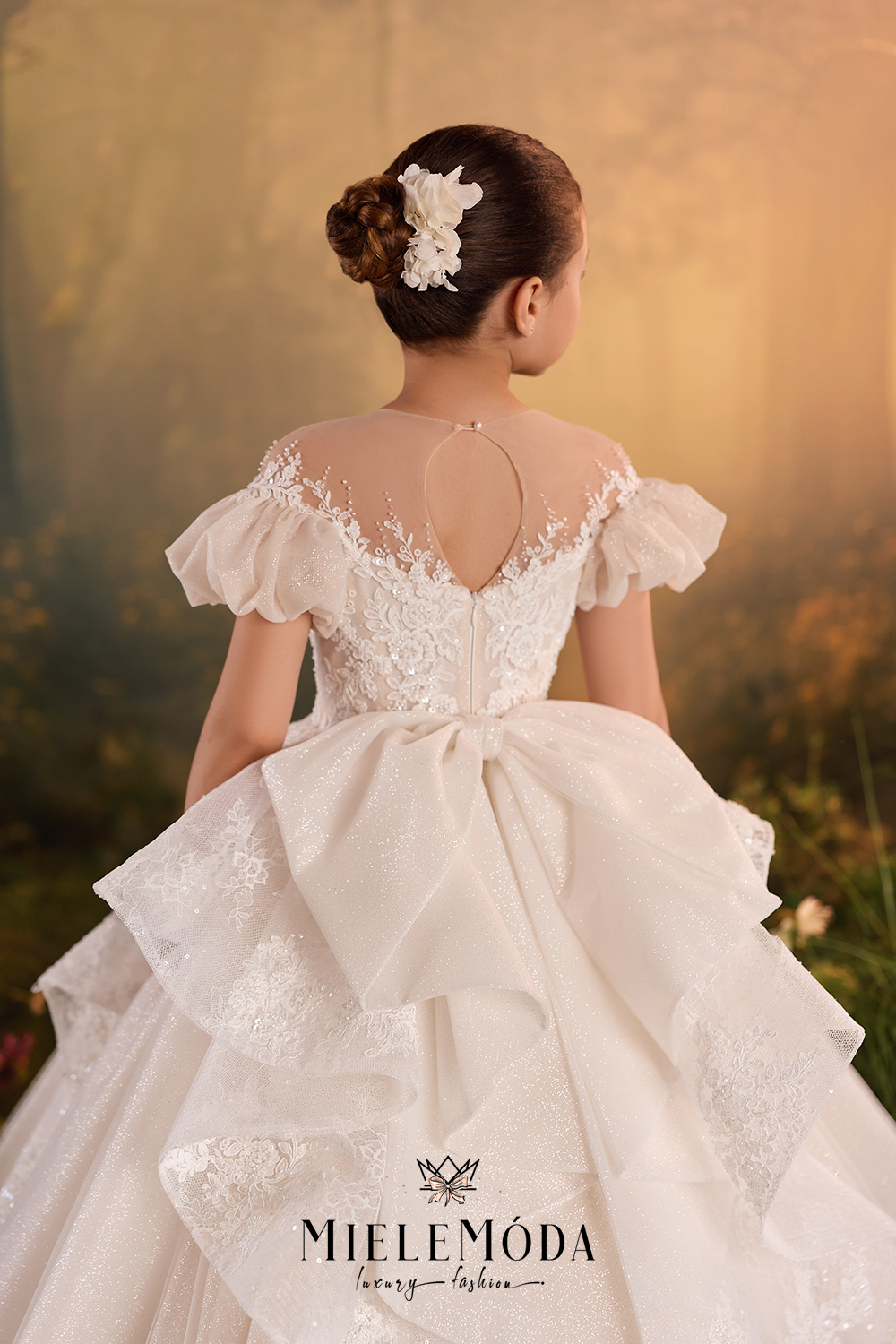 A young girl viewed from behind wearing a MieleMóda ball gown, featuring a lace bodice, ruffled off-the-shoulder sleeves, a large bow at the waist, and a layered tulle skirt. She has a white floral hair accessory in her hair, set against a warm, soft-focus outdoor background.