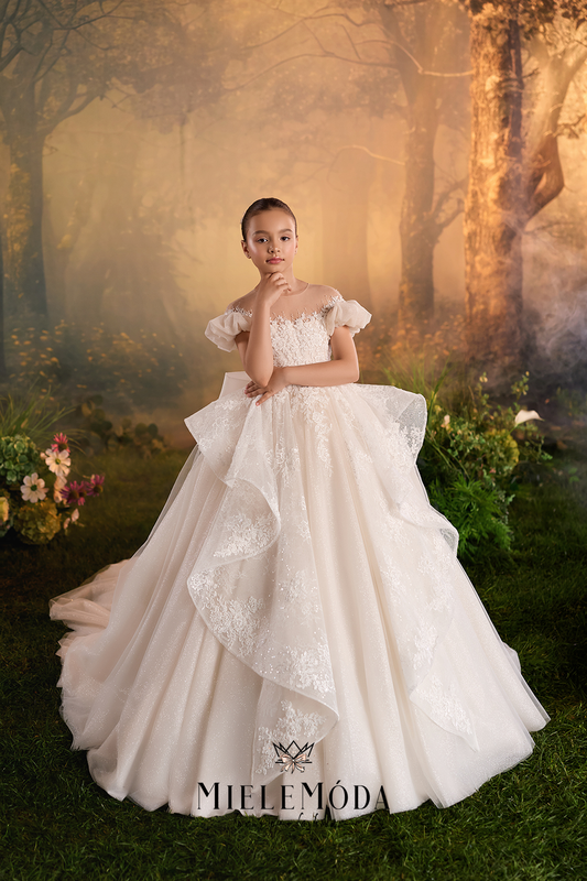 A young girl modeling a voluminous off-the-shoulder white wedding gown with lace applique and a layered ruffled overskirt, posed in a misty, golden forest setting.