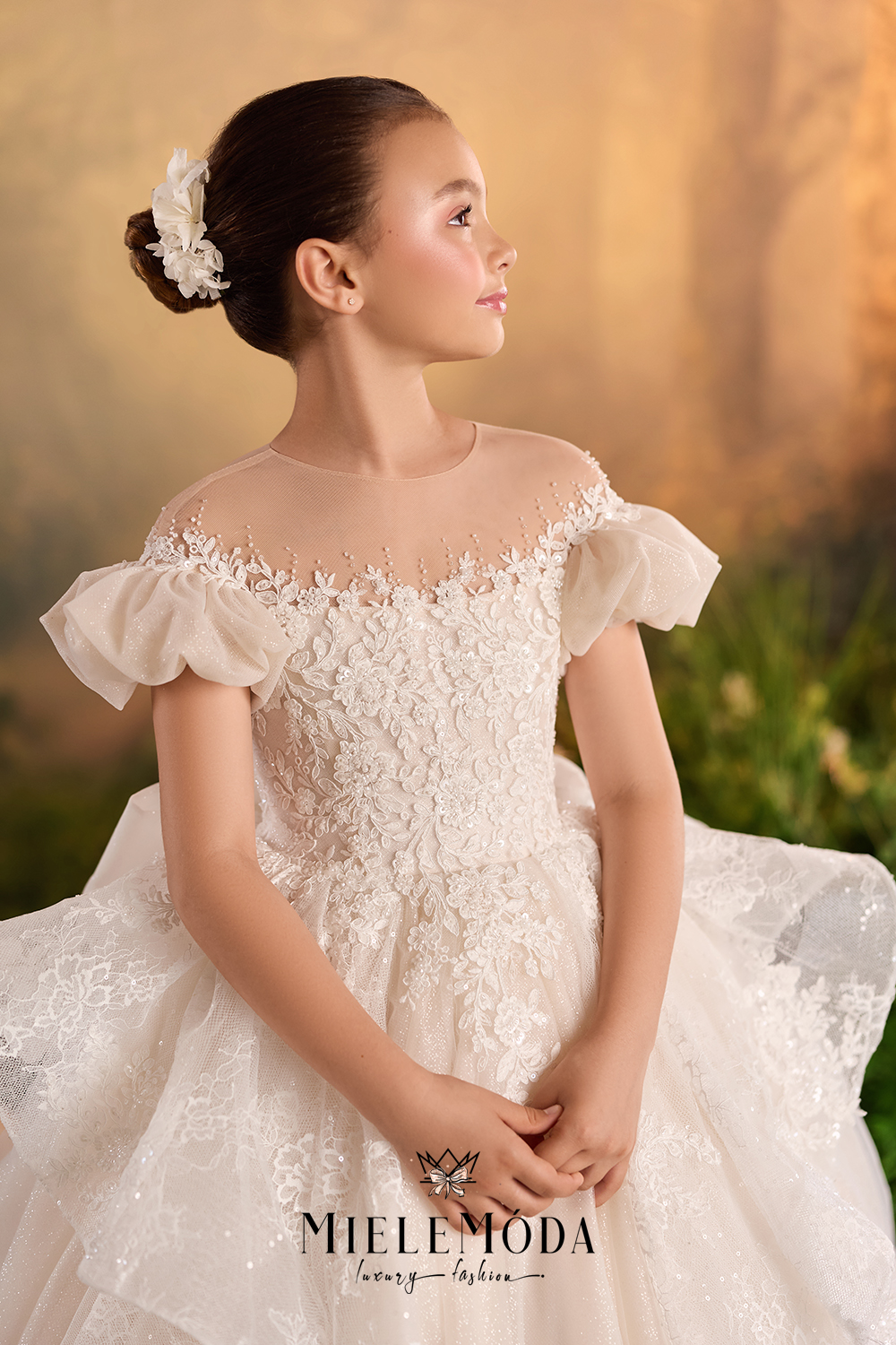 A young girl wearing a formal off-the-shoulder lace ball gown with sheer floral embroidery and a matching veil, posed against a warm golden outdoor backdrop. The brand name MieleModa is visible at the bottom.