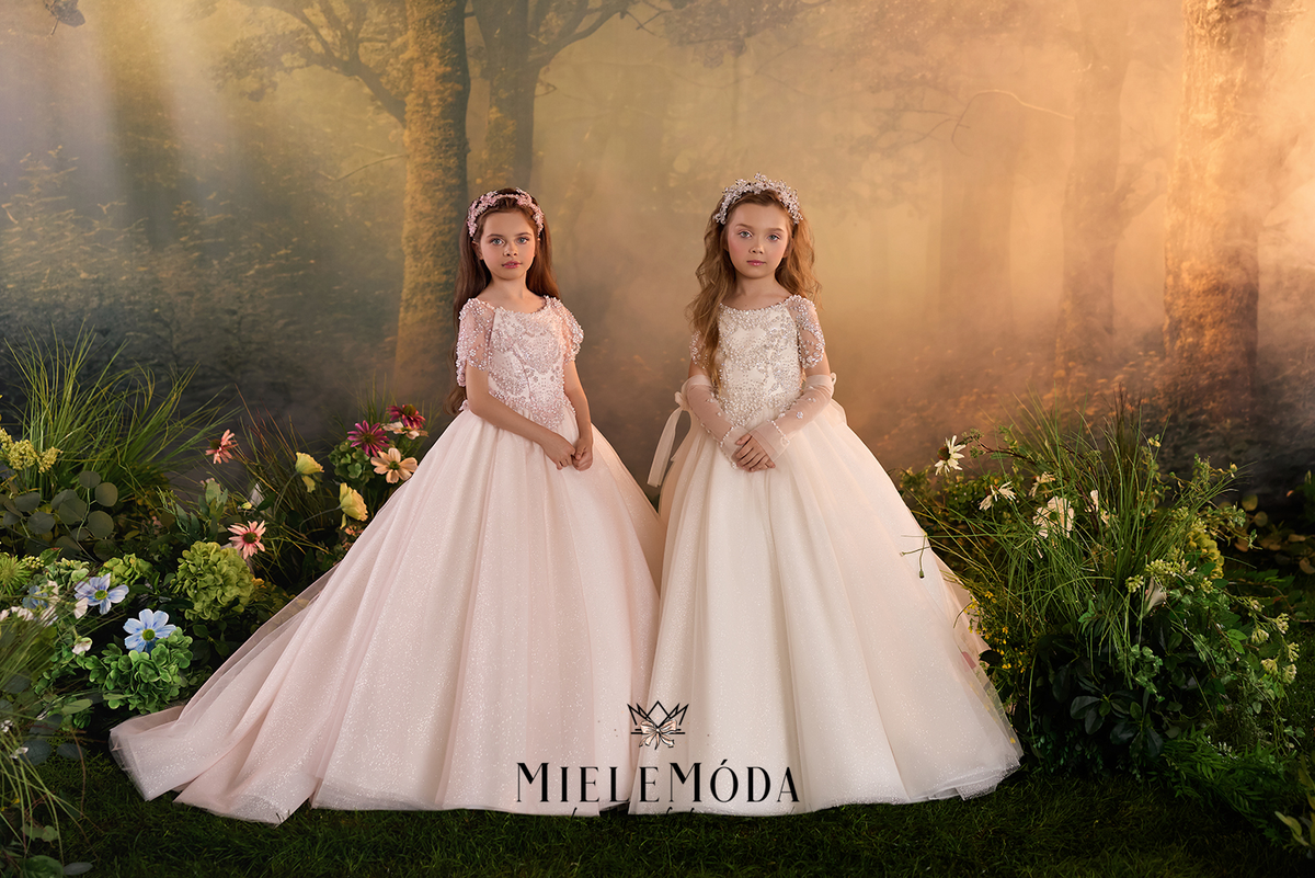 Two young girls wearing elegant, voluminous first communion dresses in pale blush pink and cream, standing in a magical, sunlit forest setting with flowers and soft mist. The brand name 'MIELEMÓDA' is displayed at the bottom.