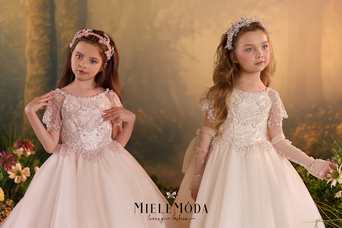 Two young girls wearing matching formal ball gowns with floral lace bodices, sheer sleeves, and full tulle skirts, posed against a soft golden forest backdrop with flowers at the sides.
