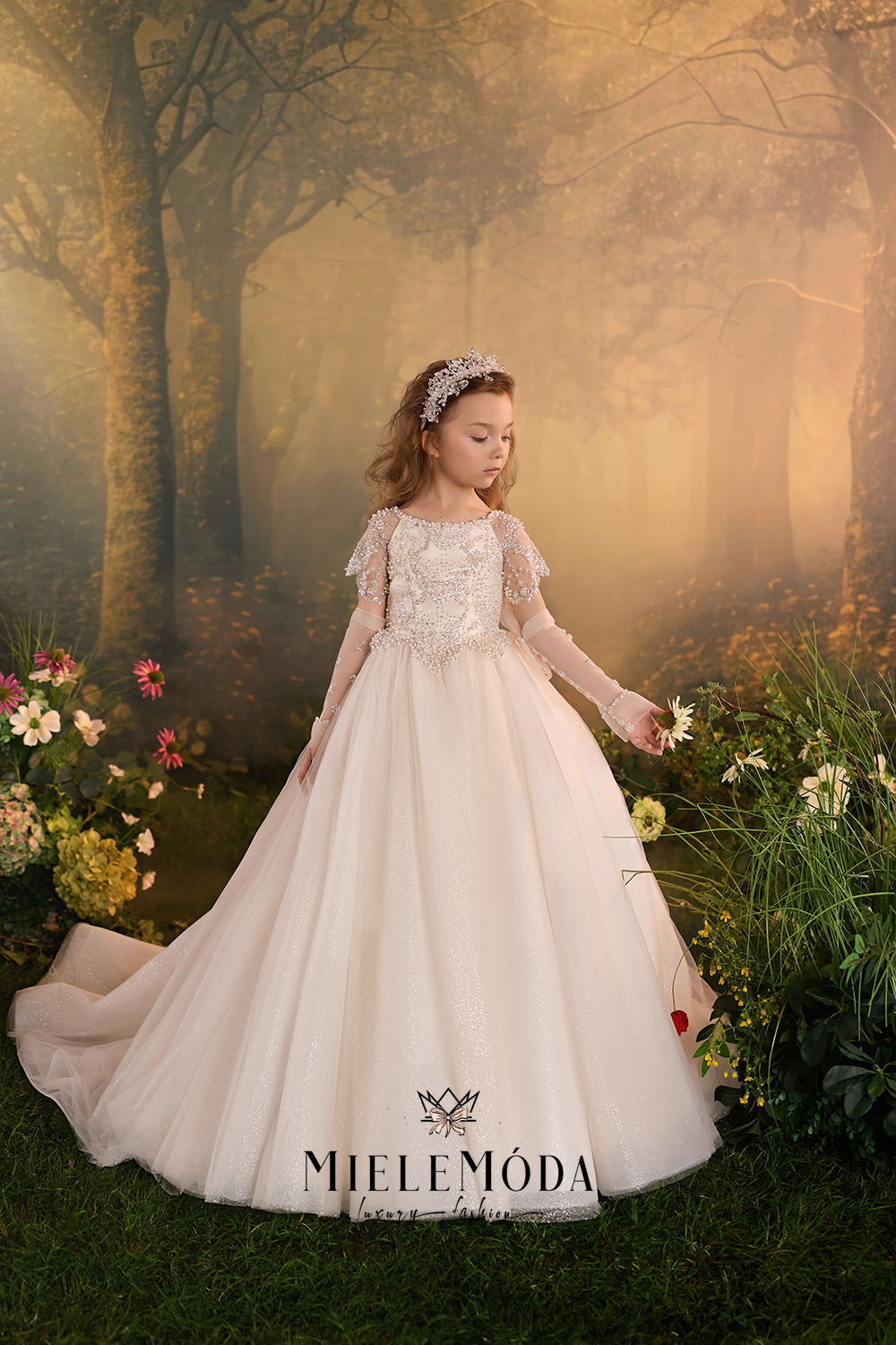 A young girl wearing a MieleMóda first communion dress stands in a misty, golden forest garden. The dress is a long, voluminous champagne-colored gown with a lace bodice, short lace sleeves, and a full tulle skirt.