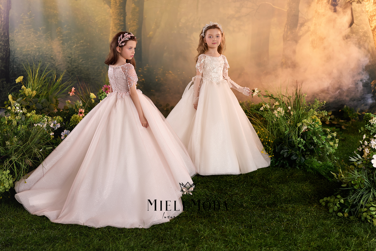 Two young girls wearing elegant off-white ball gowns with lace bodices and full tulle skirts, posed outdoors on grass with a forest backdrop and floral arrangements. The watermark 'Pappy Girl' is visible at the bottom.