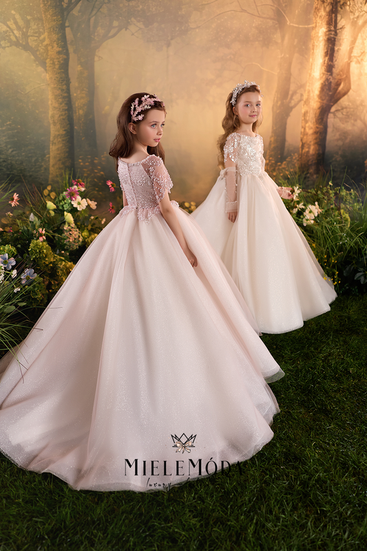 Two young girls wearing formal ball gowns in a fantasy forest setting. The girl in the foreground wears a blush pink gown with a lace bodice and short puff sleeves, while the girl behind her wears a champagne-colored gown with a lace bodice and long sleeves.