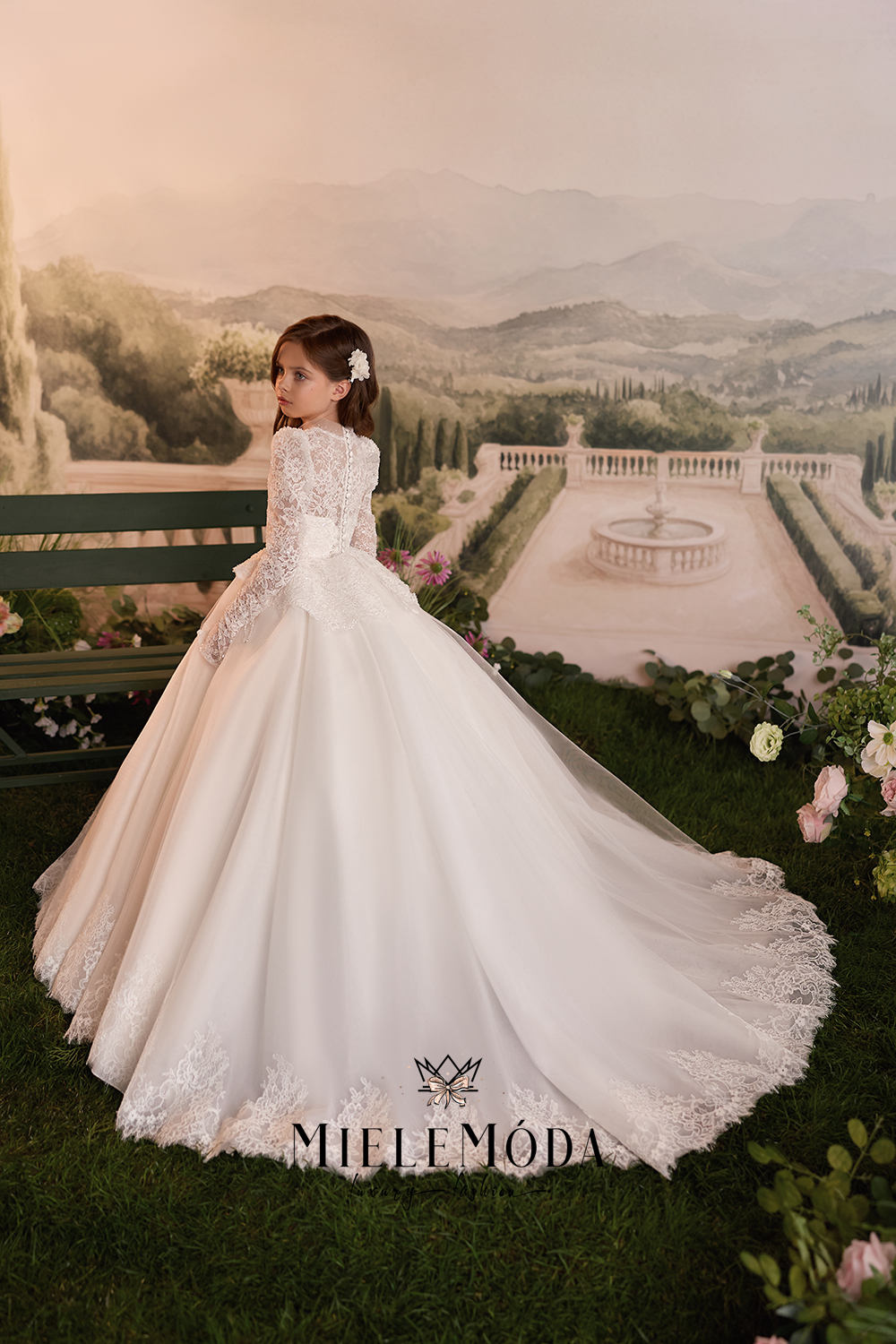 A young girl wearing a voluminous white ball gown with lace detailing and a long train, posed in front of a scenic garden backdrop featuring a fountain and distant hills.