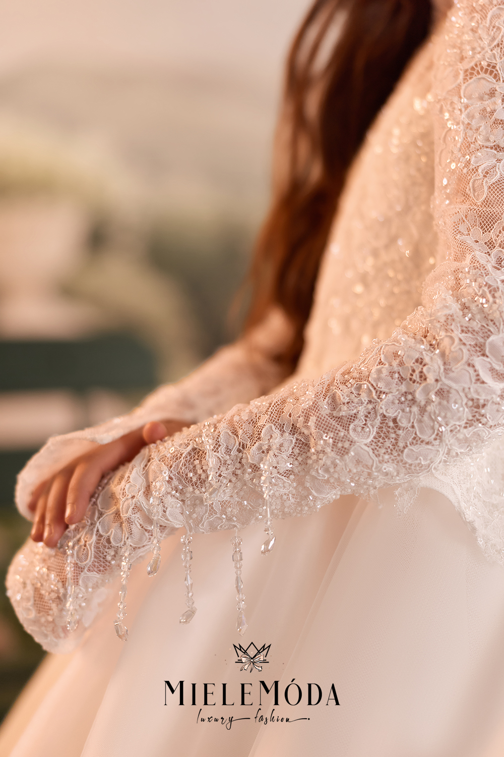 Close-up of a girl wearing a bridal gown with long sheer lace sleeves and dangling crystal embellishments, photographed in soft warm light. The MIELEMÓDA luxury fashion logo appears at the bottom.