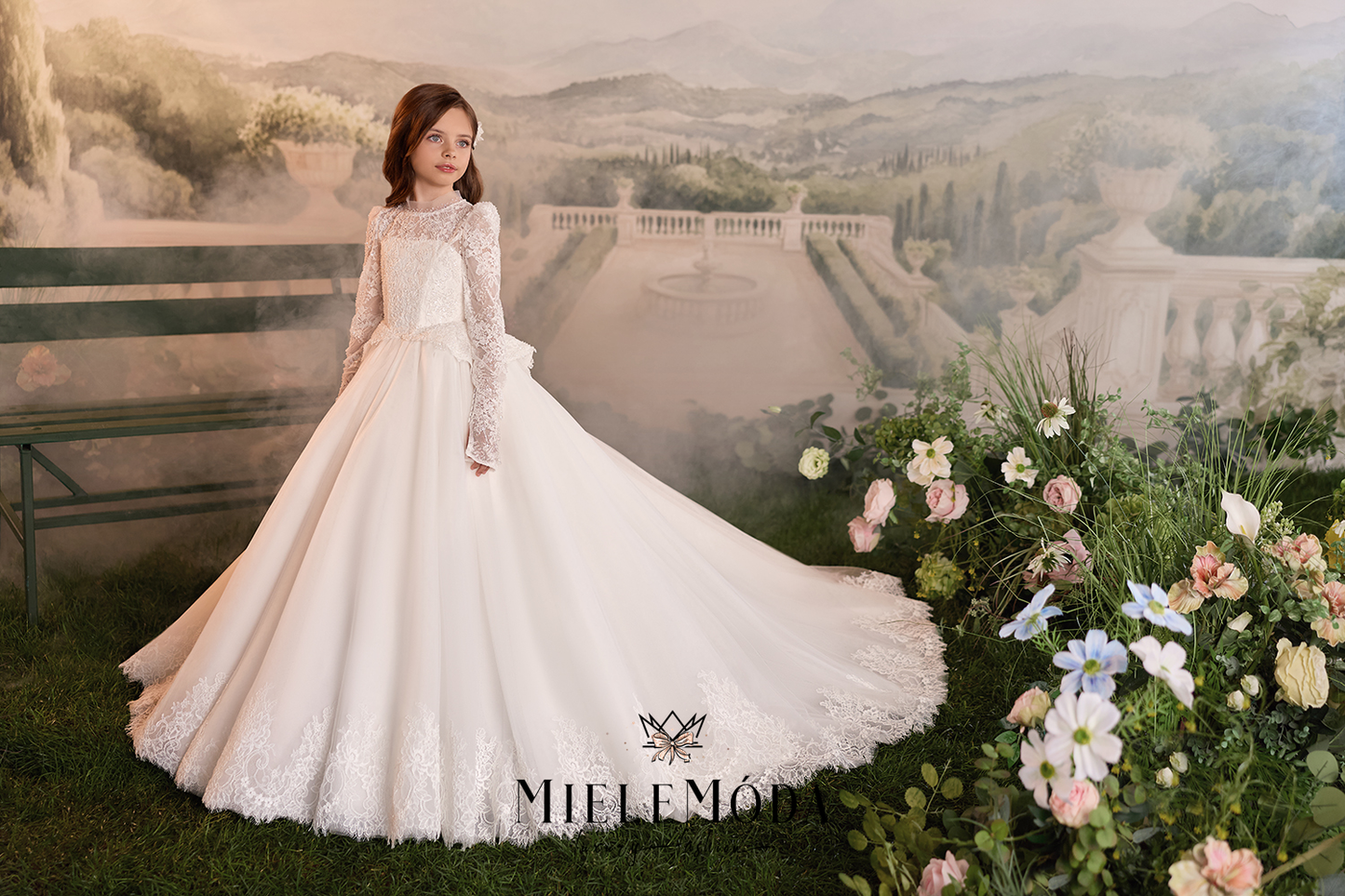 A woman wearing a voluminous white wedding gown with long lace sleeves stands in front of a scenic painted backdrop of a villa and hills, with a floral arrangement in the foreground. The brand name MIELEMÓTIS is visible at the bottom.
