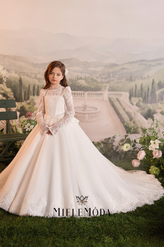 A young girl wearing a long-sleeved white ball gown with lace detailing and a full skirt, posed in front of a scenic garden backdrop. The brand name MIELEMODA is visible at the bottom.
