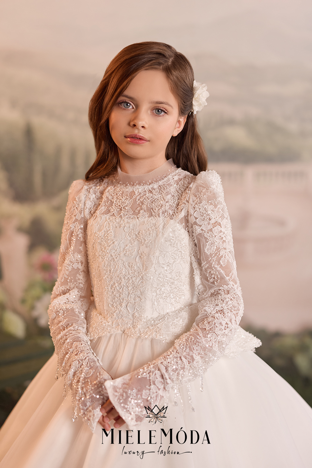 A young girl with brown hair wearing a long-sleeved white lace and tulle formal dress, posed against a soft, blurred scenic background.