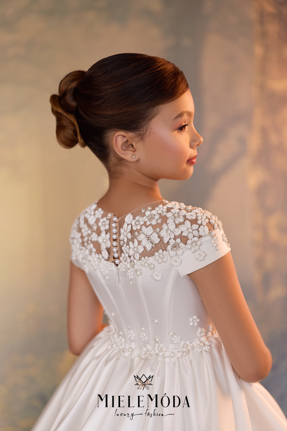 A young girl with her hair in a neat bun is shown from the back wearing a white formal dress with floral lace detailing on the bodice and short sleeves. The brand name MIELEMODA is visible at the bottom of the image.