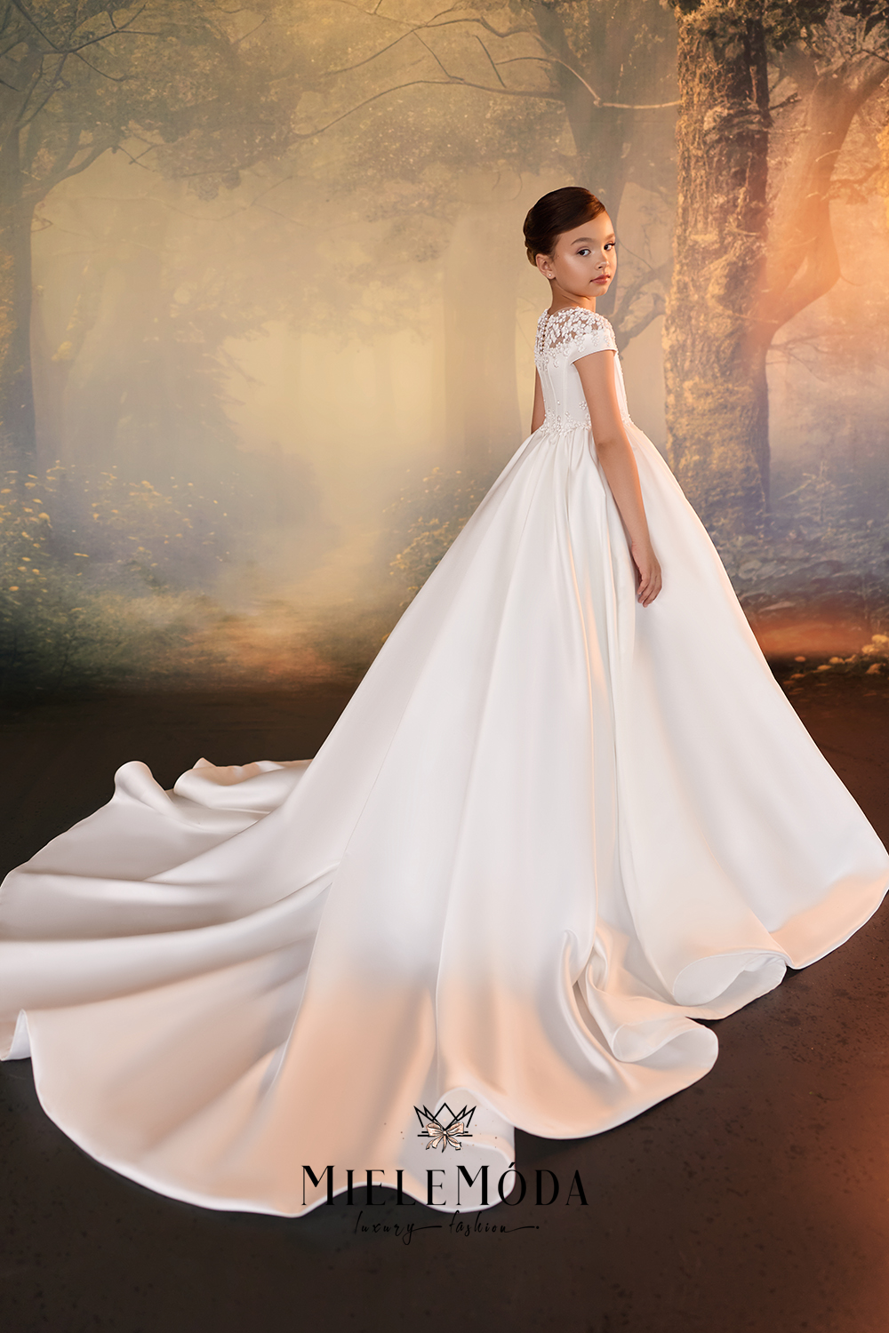 A young girl modeling a white A-line wedding dress with a short lace-embroidered bodice and a long flowing train, posed in a misty, sunlit forest setting. The brand name 'MIELEMODA' is visible at the bottom.