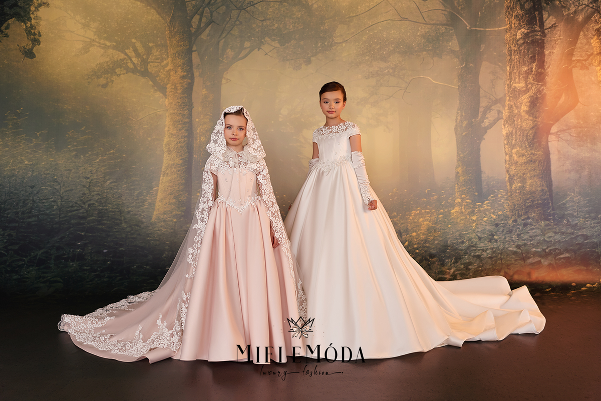 Two young girls wearing elegant ballgown wedding dresses stand in front of a misty forest backdrop. The girl on the left wears a pale blush pink gown with a matching lace-trimmed veil, and the girl on the right wears a soft ivory gown with off-the-shoulder lace sleeves.