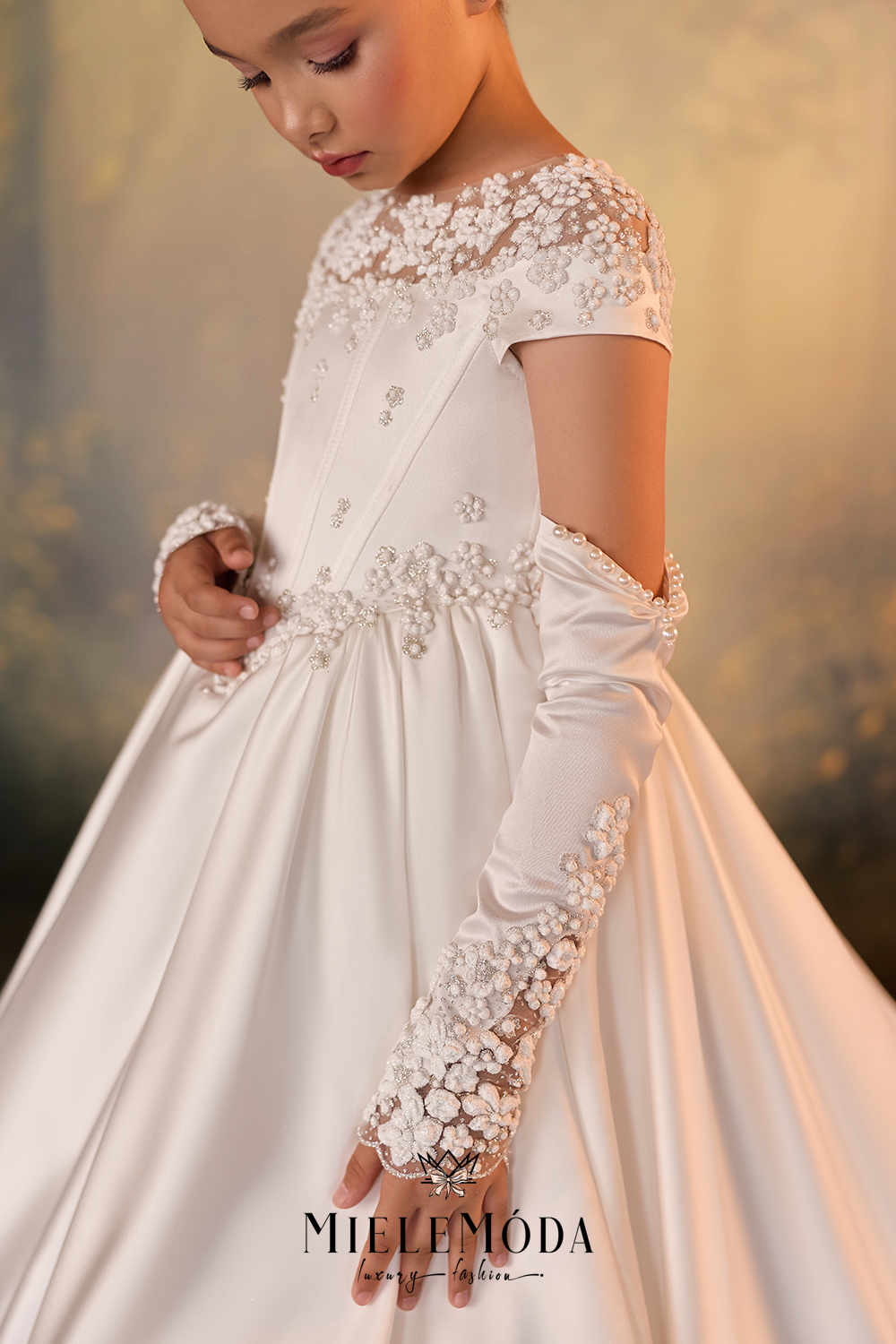 A young girl wearing a white satin ball gown with intricate floral lace and bead embellishments, paired with matching long lace gloves. The image features a warm, golden-toned blurred background and the brand name 'MILEMODA' at the bottom.