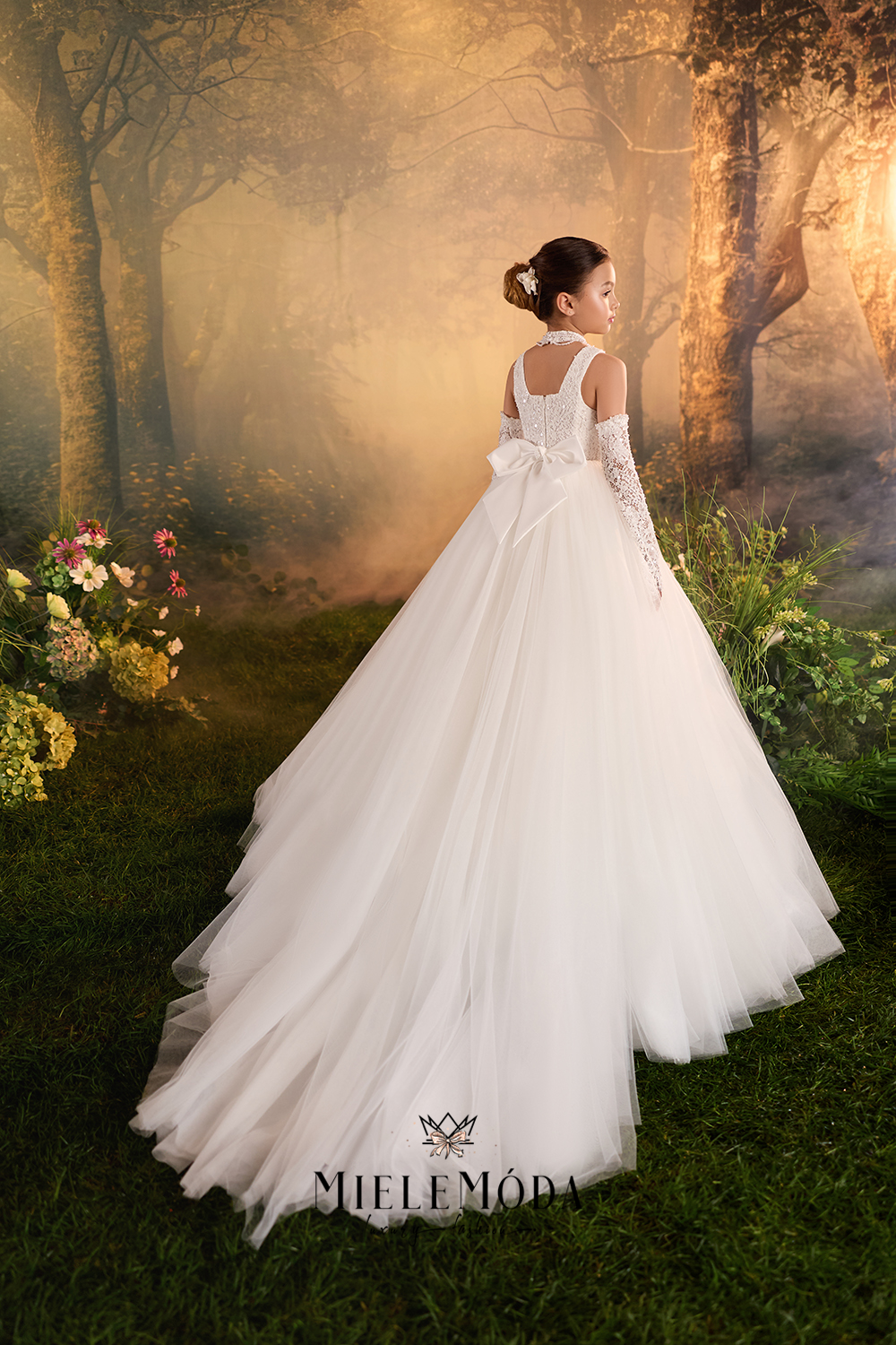 A woman wearing a white wedding gown stands in a misty forest setting, photographed from the back. The dress has long lace sleeves, a fitted bodice, and a full layered tulle skirt with a bow at the back.