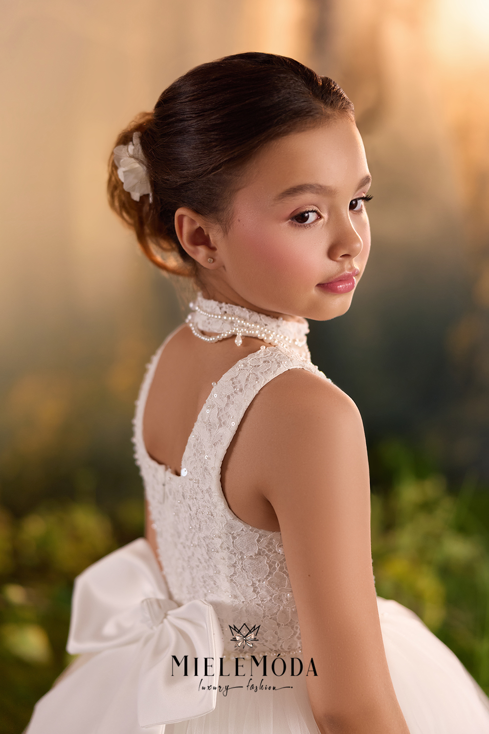 A young girl with her hair in a bun, wearing a white lace dress with a high neck, sleeveless straps, and a large bow at the waist, photographed outdoors against a warm, blurred background.