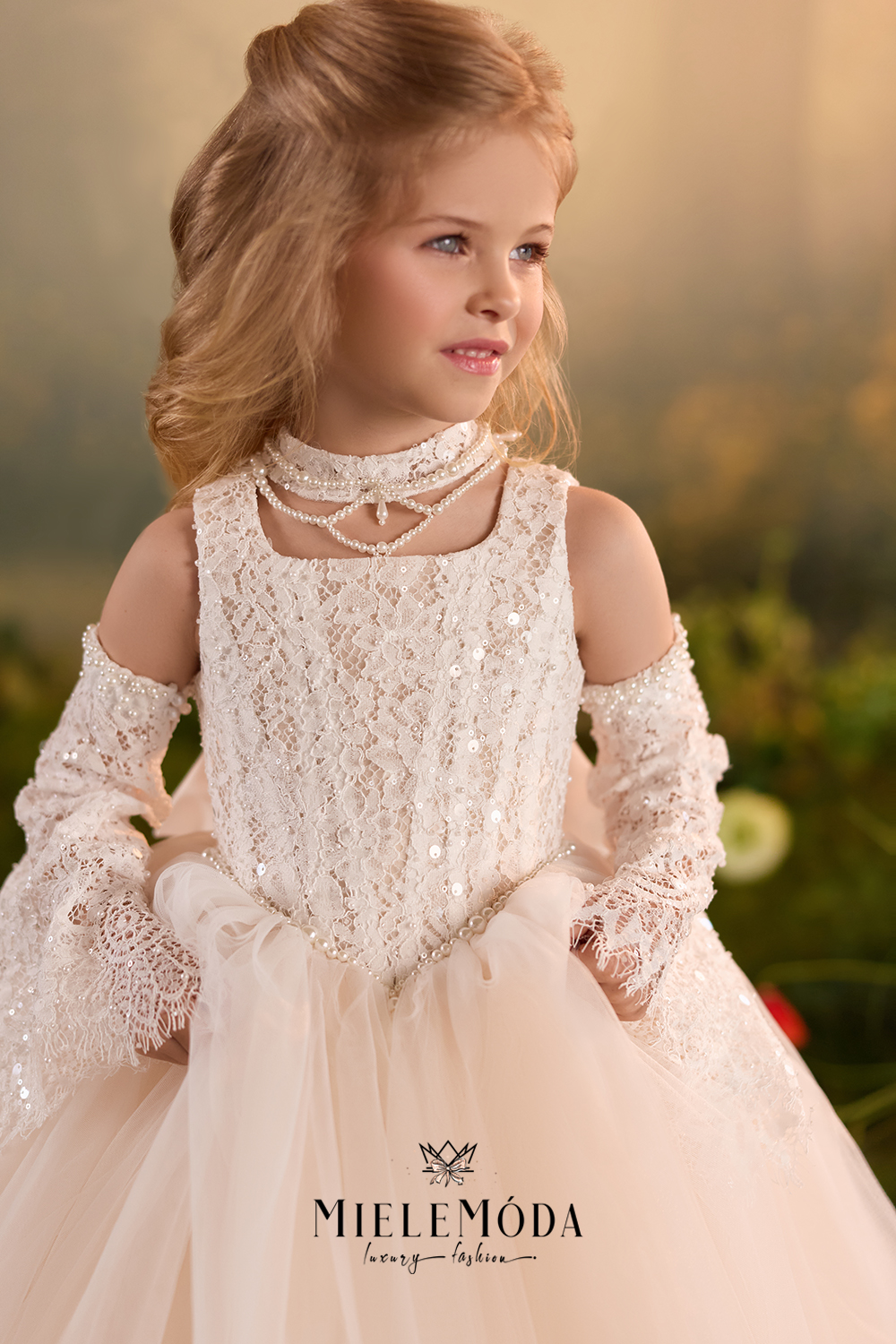 A young girl wearing a MieleMóda formal dress with a lace bodice, cold-shoulder long lace sleeves, a pearl choker, and a voluminous tulle skirt, set against a soft-focus outdoor background.