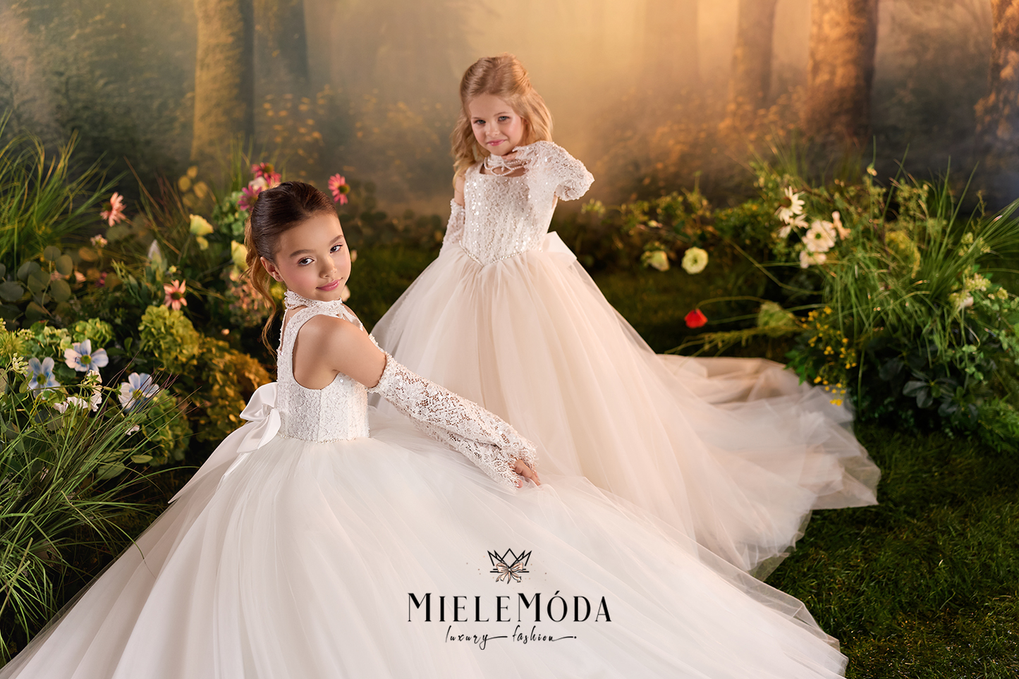 Two young girls wearing elegant white first communion dresses with lace details and long lace gloves, posed in a lush, sunlit garden setting. The brand name 'MieleMóda' is displayed at the bottom.