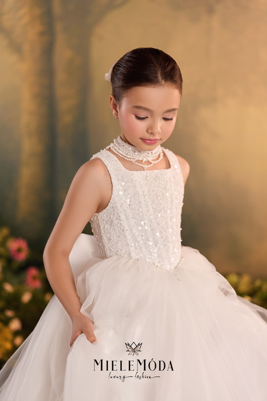 A young girl wearing a sleeveless white tulle dress with a beaded bodice and a high lace collar, posed against a warm golden floral backdrop. The brand name MieleMóda is visible at the bottom.