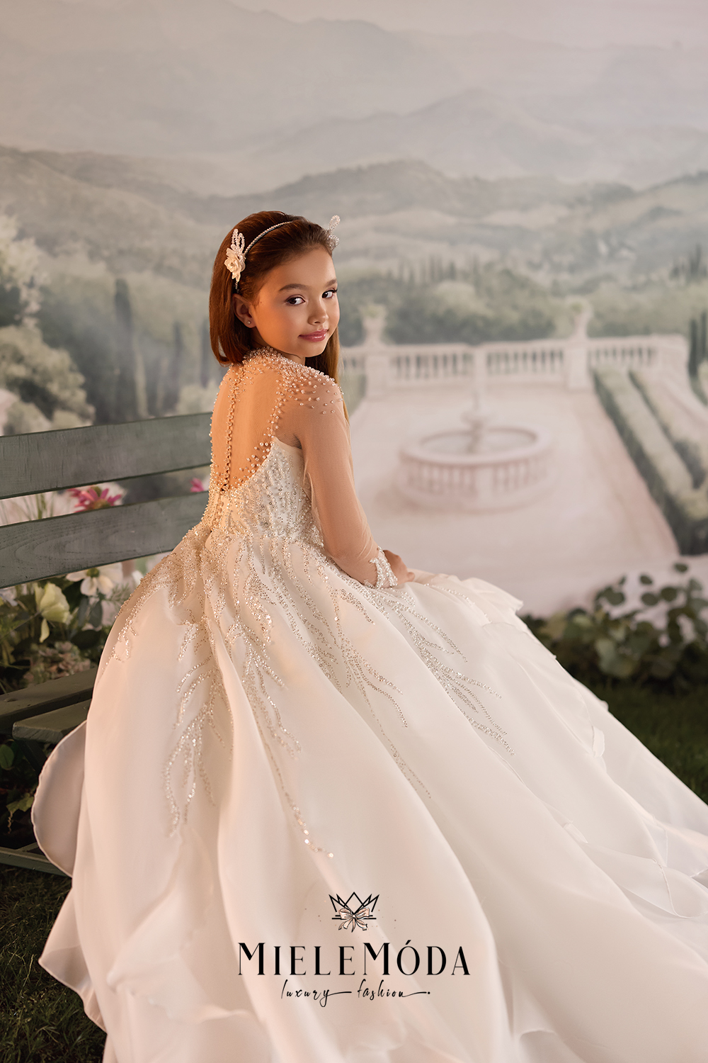 A young girl wearing a voluminous white ball gown with lace and bead embroidery, seated on a bench in front of a scenic garden backdrop.