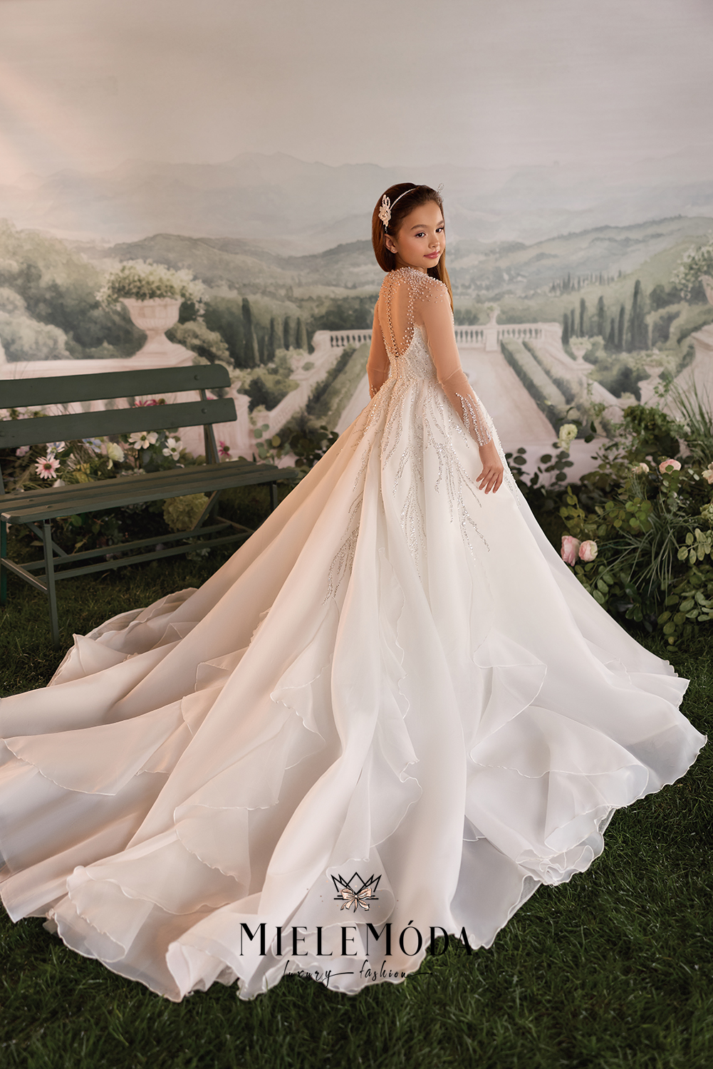 A young girl wearing a long-sleeve ivory wedding dress with lace detailing and a full train, standing in front of a scenic garden backdrop. The brand name 'MIELEMÓDA' is visible at the bottom.