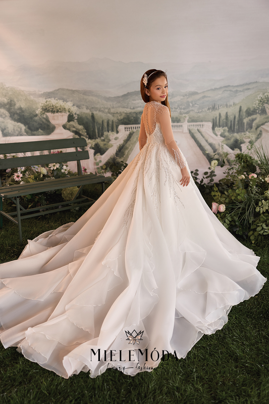 A young girl wearing a long-sleeve ivory wedding dress with lace detailing and a full train, standing in front of a scenic garden backdrop. The brand name 'MIELEMÓDA' is visible at the bottom.