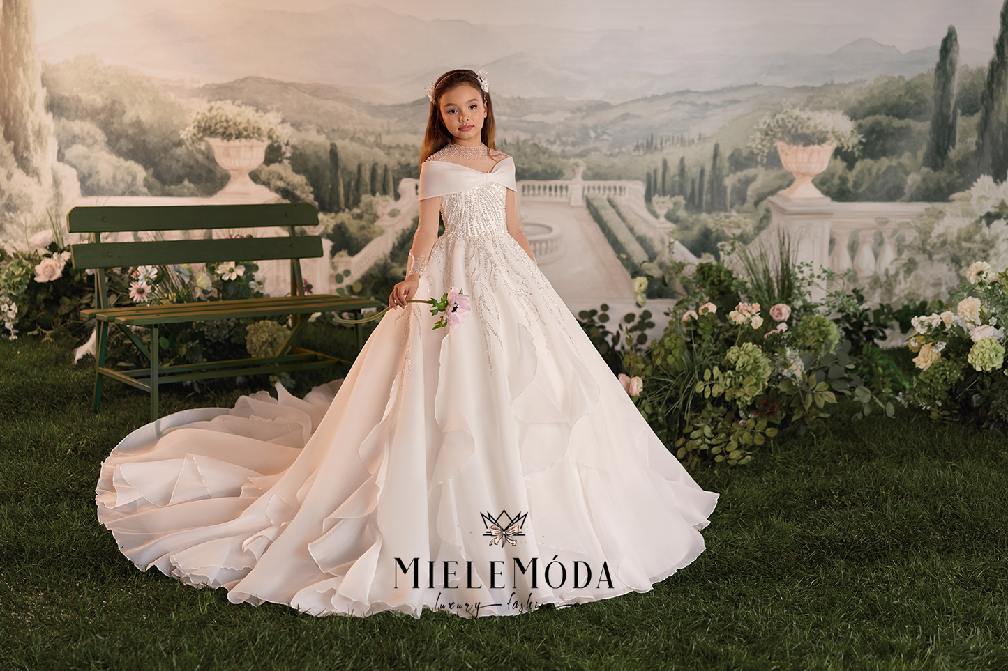 A young girl models a Miele Móda off-the-shoulder ball gown wedding dress in front of a scenic garden backdrop featuring a bench, flowers, and a path leading to a distant villa.