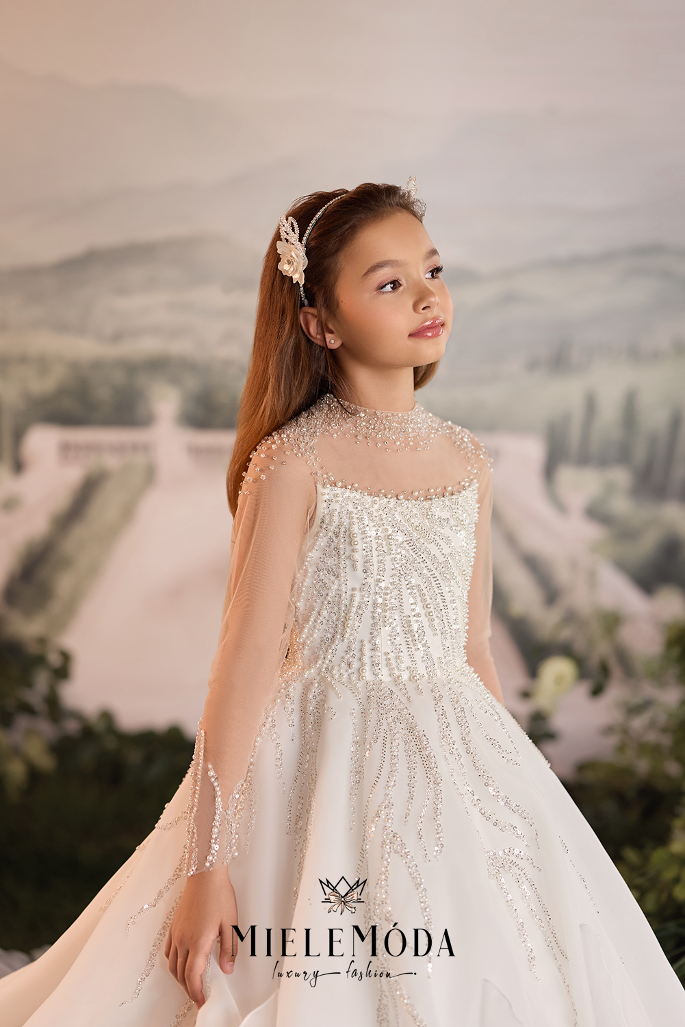 A young girl wearing a white beaded ball gown with sheer long sleeves and a floral headpiece, posed against a soft, scenic background. The brand name 'MIELEMODA' is visible at the bottom.