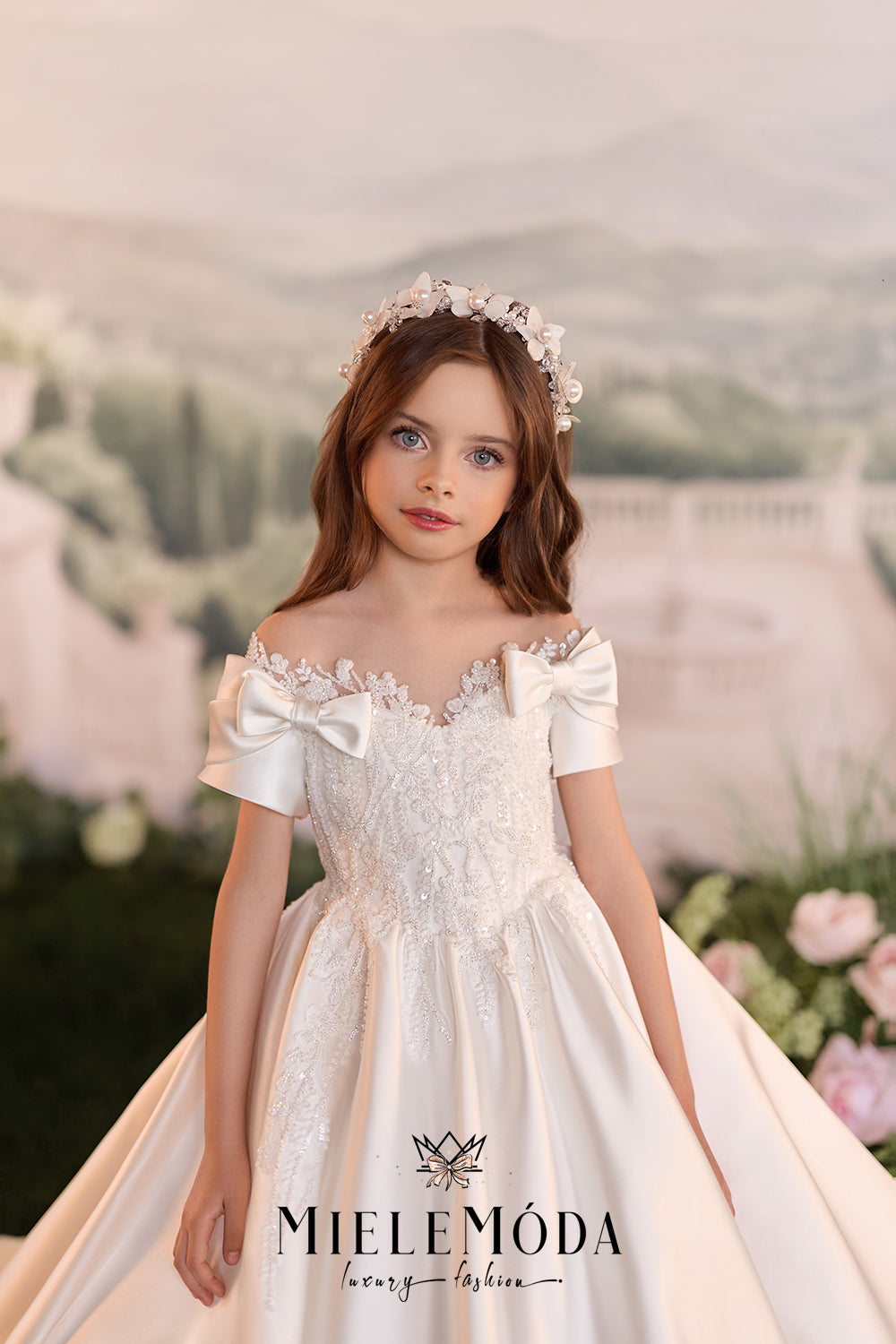 A young girl wearing a formal off-the-shoulder white lace ball gown with large bow details, a matching floral crown, and a voluminous skirt, posed against a soft blurred scenic background. The brand name MIELEMODA is visible at the bottom.
