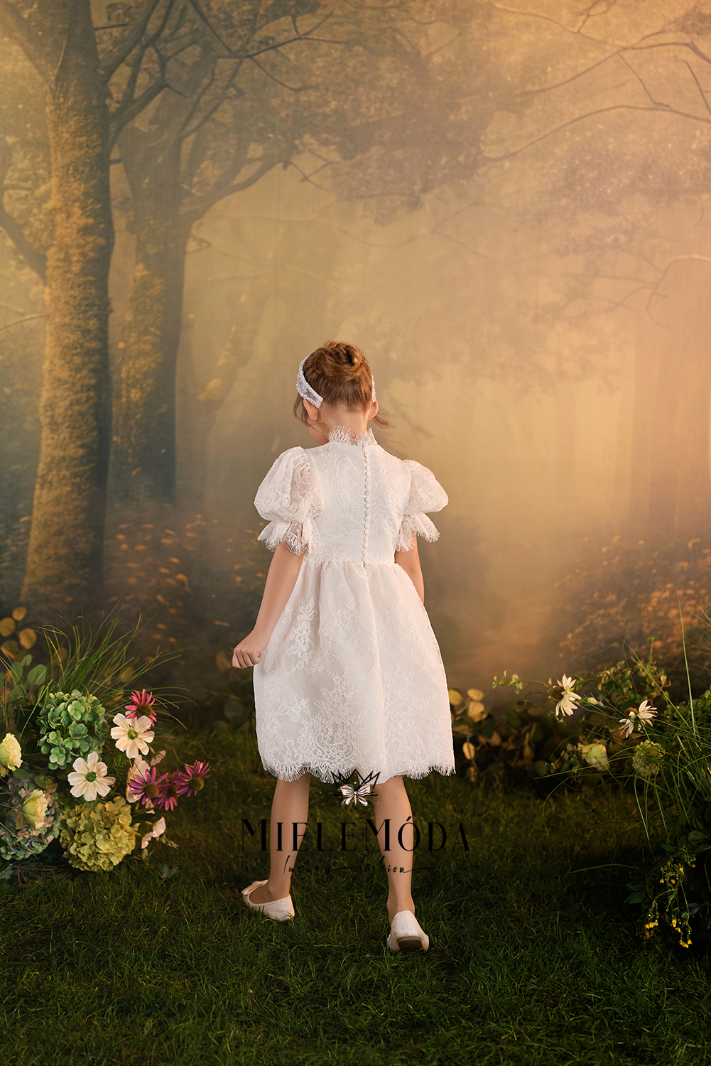 A young girl viewed from behind wearing a white lace dress and white shoes, standing in a misty, golden woodland scene with flowers at her sides.