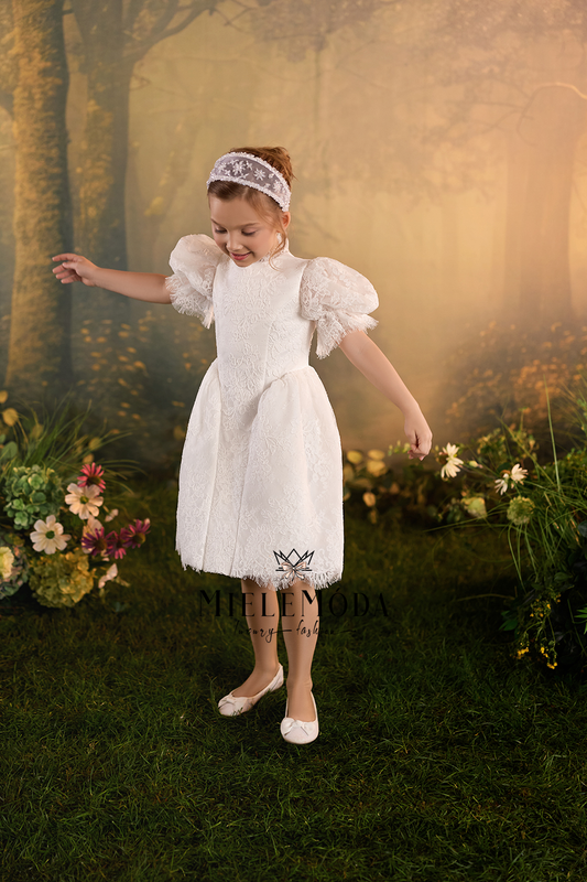A young girl wearing a white lace flower girl dress with puff sleeves, a matching white lace headband, and white shoes, posing in a misty, golden forest setting.