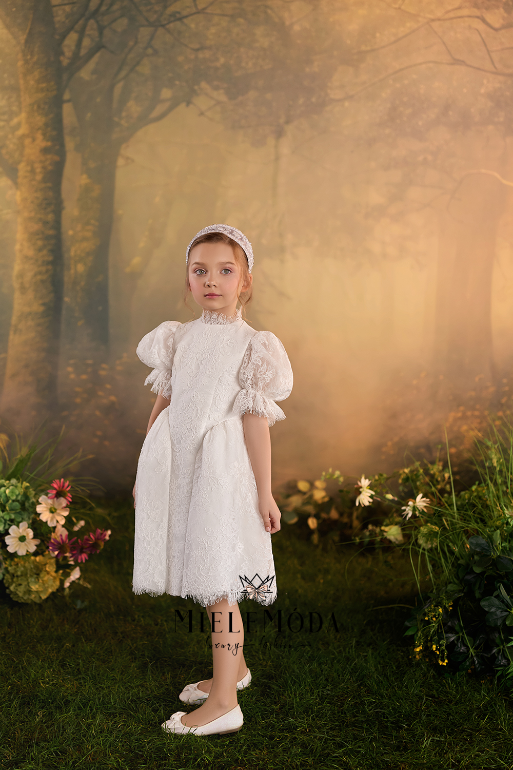 A young girl stands in a misty, golden-toned forest setting wearing a white lace and textured dress with puffed sleeves, a matching headband, and white flats. Floral arrangements line the sides of the scene.