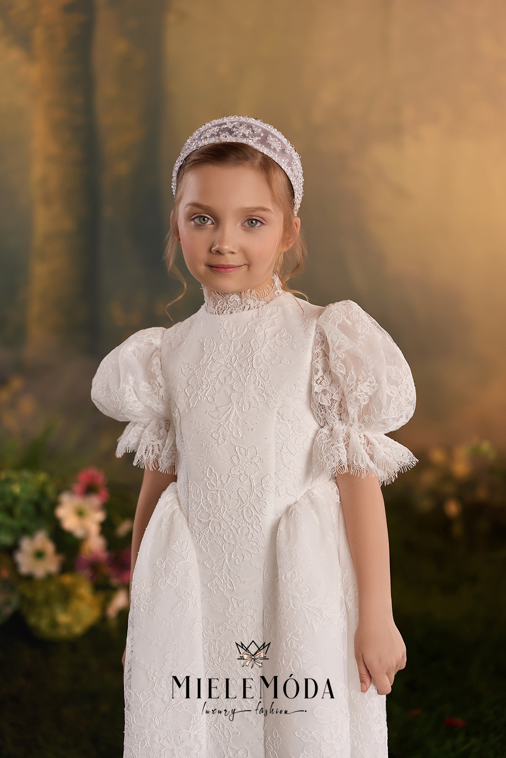 A young girl wearing a white lace dress with puff sleeves, a high ruffled collar, and a matching lace headband, posed against a warm, blurred forest background. The brand name 'MIELEMÓDA' is printed on the dress.