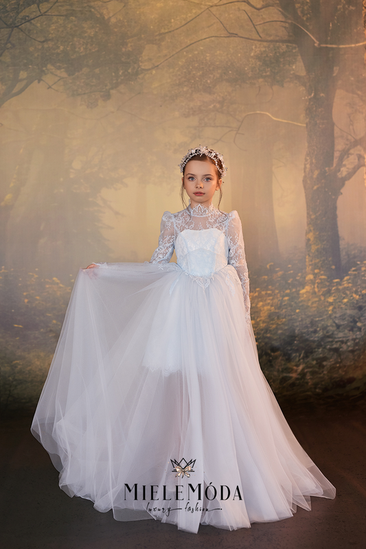 A young girl wearing a long-sleeved white lace and tulle ball gown with a sweetheart bodice, holding out the skirt to show its volume, set against a misty forest background. The brand name 'MIELEMODA' is visible at the bottom.