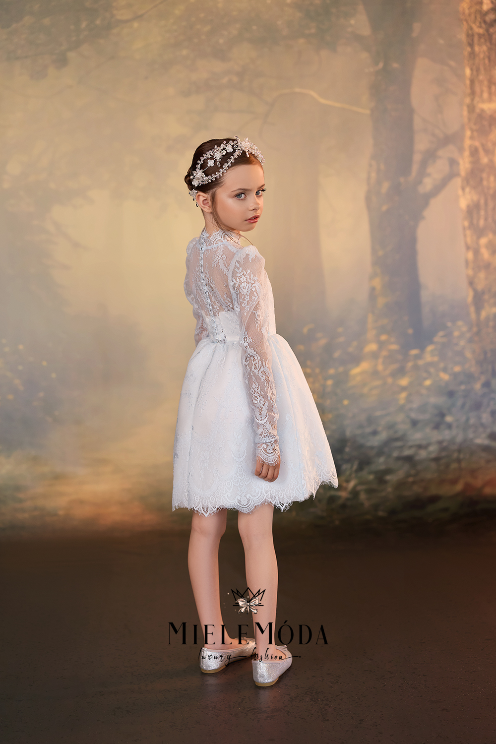 A young girl stands in profile wearing a white lace and tulle flower girl dress with silver shoes, set against a soft, misty forest backdrop. The brand name 'Mielemóda' is visible as a watermark at the bottom.