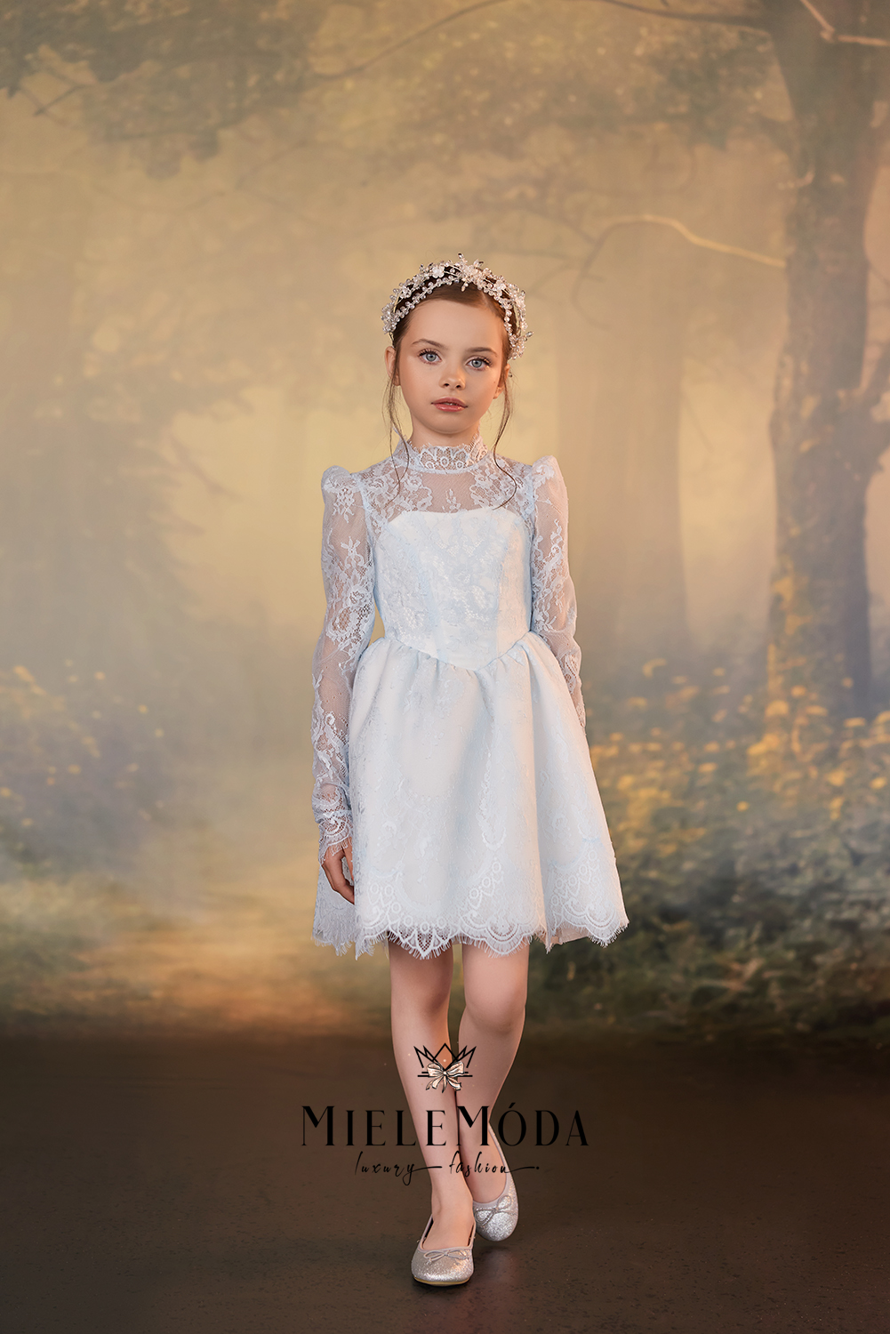 A young girl stands in a misty forest setting wearing a white first communion dress with long sheer lace sleeves, a lace-trimmed skirt, and a matching floral crown. The brand name MieleMóda is visible at the bottom of the image.