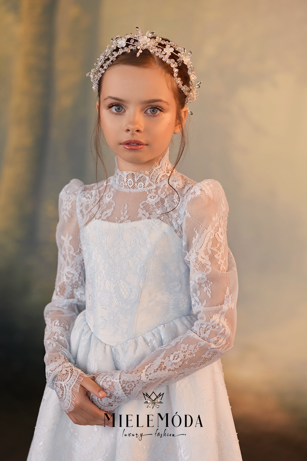 A young girl wearing a white lace dress with sheer long sleeves, a high lace collar, and a matching white crown, posed against a soft, warm-toned blurred background. The brand name 'MIELEMÓDA' is visible at the bottom.