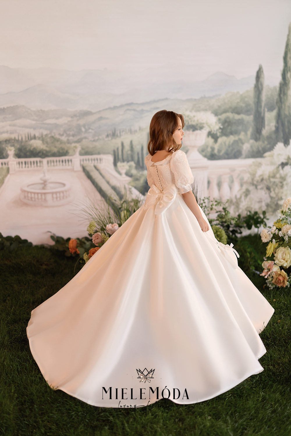 A woman wearing a white A-line wedding dress with short puff sleeves and a lace back, standing in front of a scenic garden backdrop. The brand name 'MIELEMODA' is printed on the bottom of the dress.