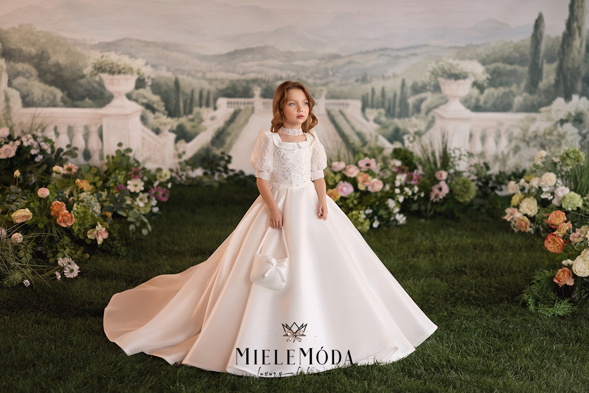 A young girl stands in a formal white ball gown with short puff sleeves and a full skirt, holding a small white handbag. The scene is set on green grass with floral arrangements and a scenic garden backdrop.