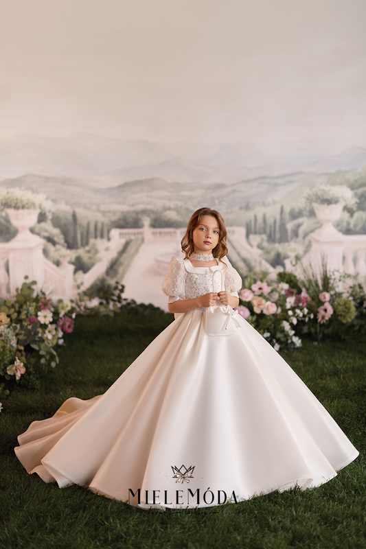 A young girl stands in a formal garden wearing a voluminous white first communion dress with a lace bodice and short sleeves. The dress has a long train spread across green grass, with a scenic garden backdrop featuring a path, hills, and floral arrangements.