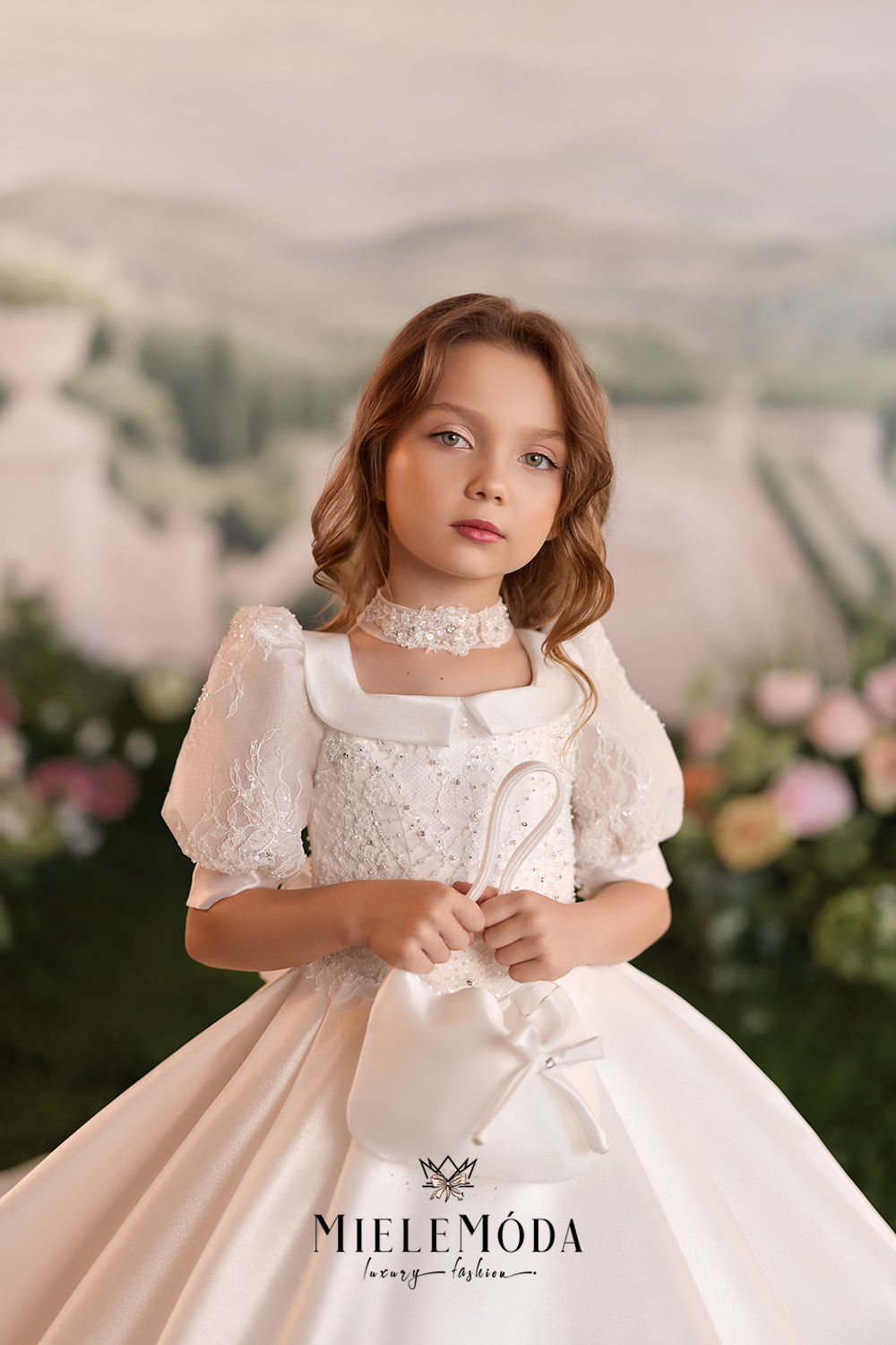 A young girl wearing a formal white lace ball gown with puff sleeves and a large bow at the waist, standing against a soft blurred garden background. The brand name MIELEŌōA is visible at the bottom.