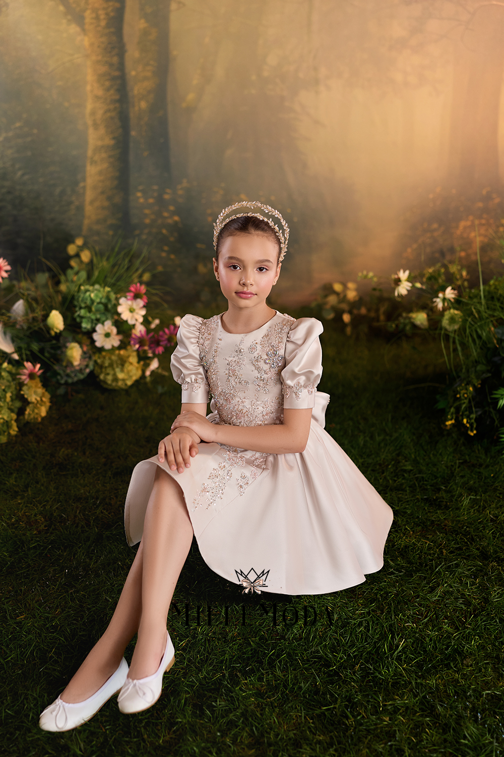 A young girl sits on grass in a whimsical, misty forest setting, wearing a formal light pink dress with puffed sleeves, a matching headpiece, and white shoes.