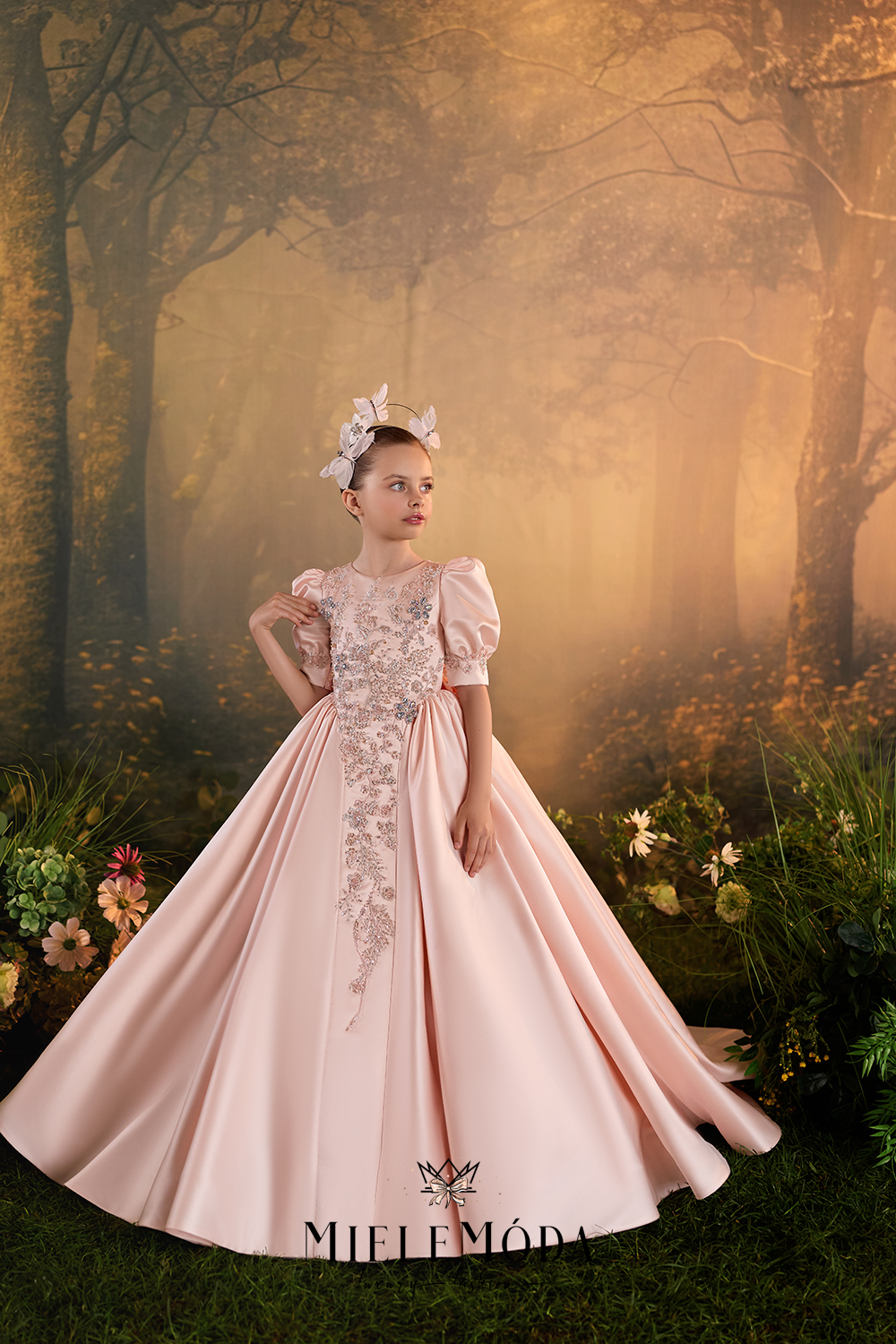 A young girl wearing a pale blush pink ball gown with lace detailing and a matching headpiece, posed in a misty, golden-toned forest setting. The brand name 'MIELEMÓD' is visible at the bottom.