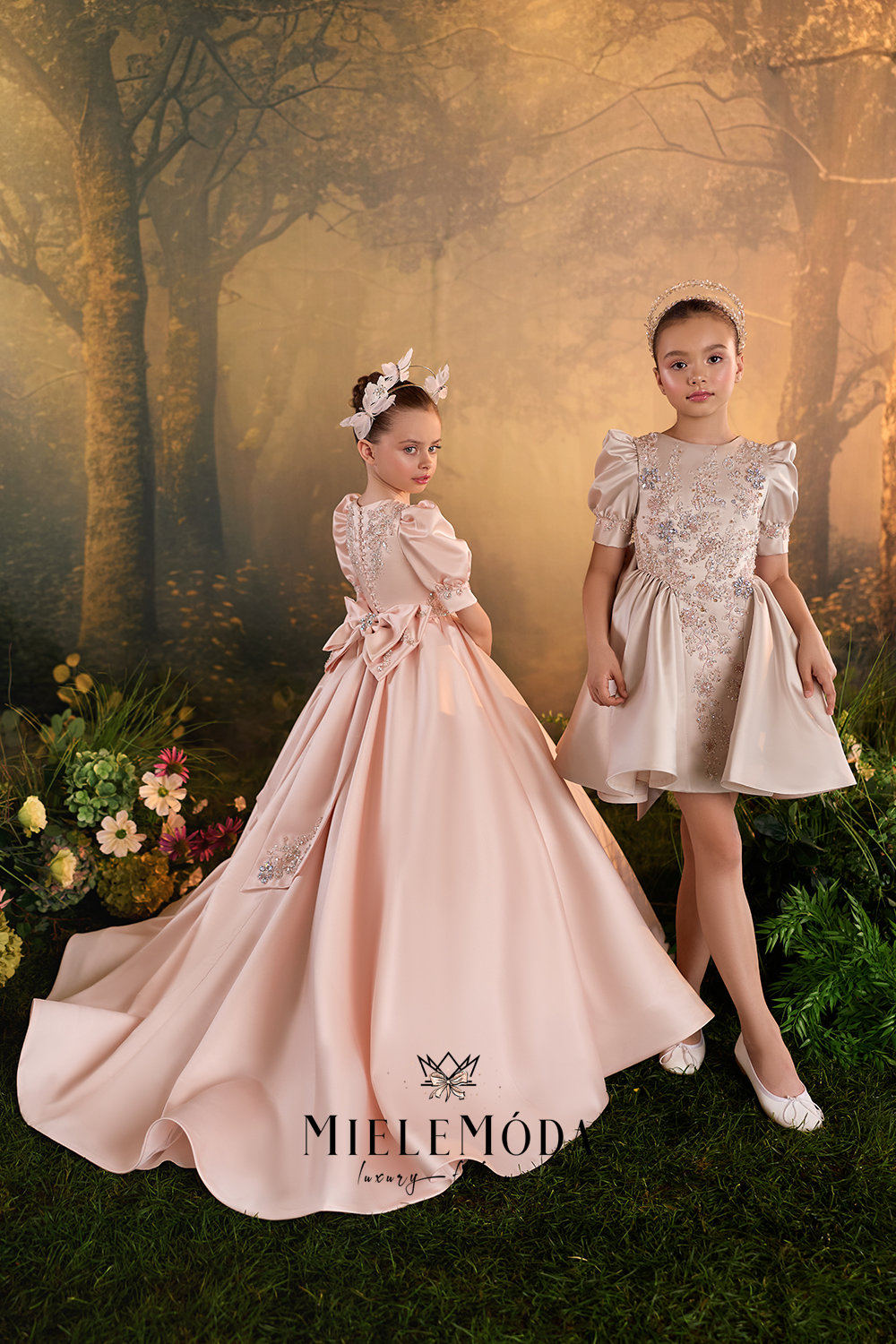 Two young girls modeling formal ballgown party dresses in a fantasy forest setting. The girl on the left wears a long, voluminous light pink gown with puffed sleeves and a bow at the waist, while the girl on the right wears a shorter champagne beige lace dress with puffed sleeves.