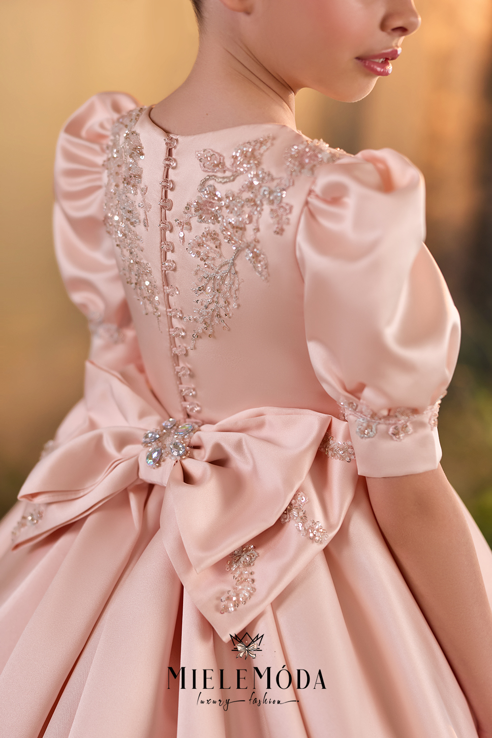 A close-up back view of a child wearing a pale blush pink ball gown with puffed sleeves, a large bow at the back, and ornate crystal embellishment. The brand name MIELEMODA is visible at the bottom.