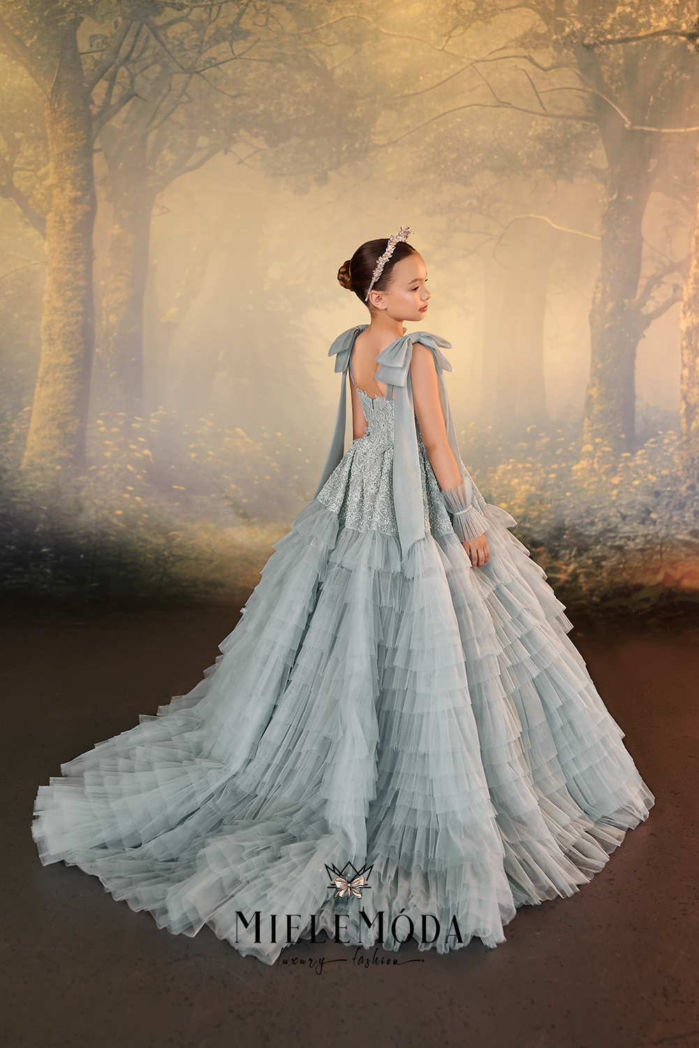 A young girl wearing a voluminous light blue tulle ball gown with a ruffled back and matching gloves, posed in a misty, golden-toned forest setting.
