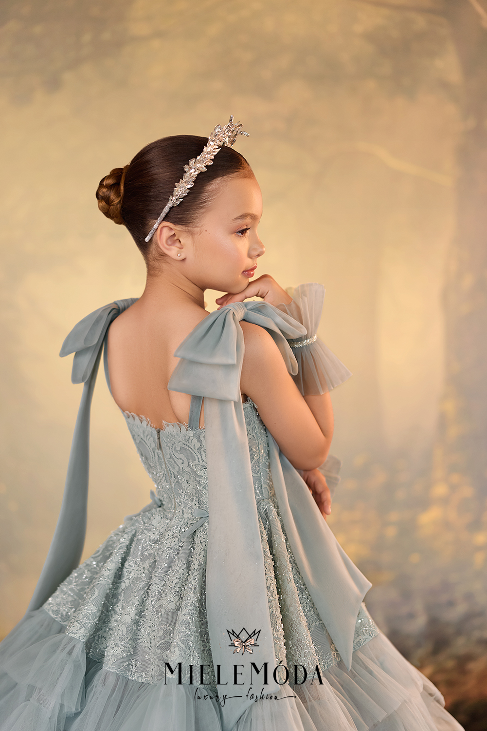 A young girl in profile wearing a light blue ball gown with large bow details on the shoulders, a matching headpiece, and a tiara, posed against a soft, warm golden background. The brand name 'MIELEMODA' is visible at the bottom.