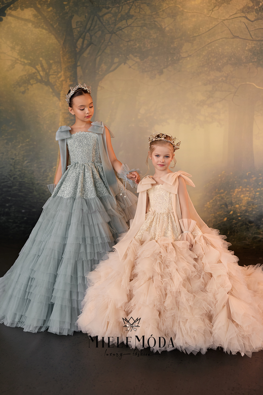 Two young girls in elaborate ball gowns with matching tiaras, posed in a misty forest setting. The girl on the left wears a light blue-gray gown with a sheer cape, and the girl on the right wears a champagne beige gown with a feathered skirt and sheer cape.